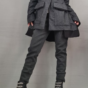 K&ouml;nnte beinhalten: Ein dunkelgrauer Blazer mit einem Knopfverschluss und gro&szlig;en Pattentaschen, getragen &uuml;ber einem wei&szlig;en Oberteil mit hohem Kragen. Das Outfit umfasst eine dunkelgraue, eng anliegende Hose und schwarze Stiefeletten mit silberfarbenen Ketten.