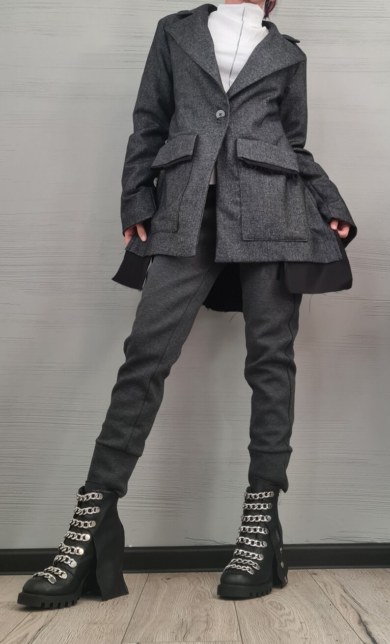 K&ouml;nnte beinhalten: Ein dunkelgrauer Blazer mit einem Knopfverschluss und gro&szlig;en Pattentaschen, getragen &uuml;ber einem wei&szlig;en Oberteil mit hohem Kragen. Das Outfit umfasst eine dunkelgraue, eng anliegende Hose und schwarze Stiefeletten mit silberfarbenen Ketten.