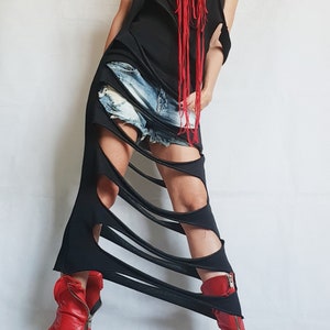 May include: A black, sleeveless dress with a shredded design, paired with denim shorts. The dress features cut-out details and is accessorized with a red fringe necklace. The outfit is completed with red leather ankle boots with zippers.
