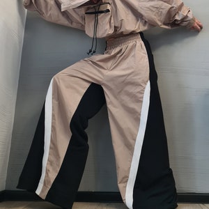 May include: A beige hooded windbreaker jacket with a front utility pocket and black drawstrings, paired with wide-leg trousers featuring a color-blocked design of beige and black panels separated by a vertical white stripe and an elastic waistband.