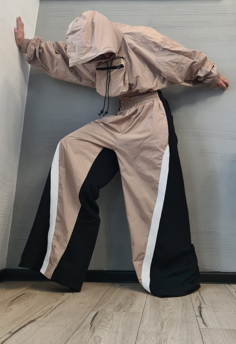 May include: A beige hooded windbreaker jacket with a front utility pocket and black drawstrings, paired with wide-leg trousers featuring a color-blocked design of beige and black panels separated by a vertical white stripe and an elastic waistband.