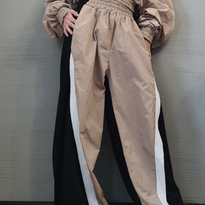May include: A beige hooded pullover with a front pouch pocket and long sleeves with ribbed cuffs, paired with high-waisted trousers featuring a wide, gathered elastic waistband. The trousers have a color-blocked design with beige and black panels.