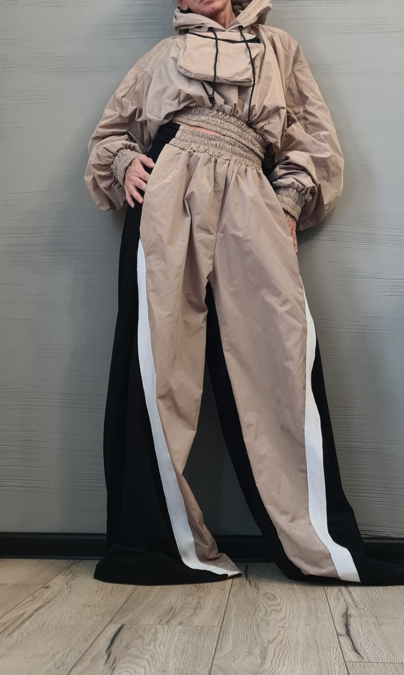 May include: A beige hooded pullover with a front pouch pocket and long sleeves with ribbed cuffs, paired with high-waisted trousers featuring a wide, gathered elastic waistband. The trousers have a color-blocked design with beige and black panels.