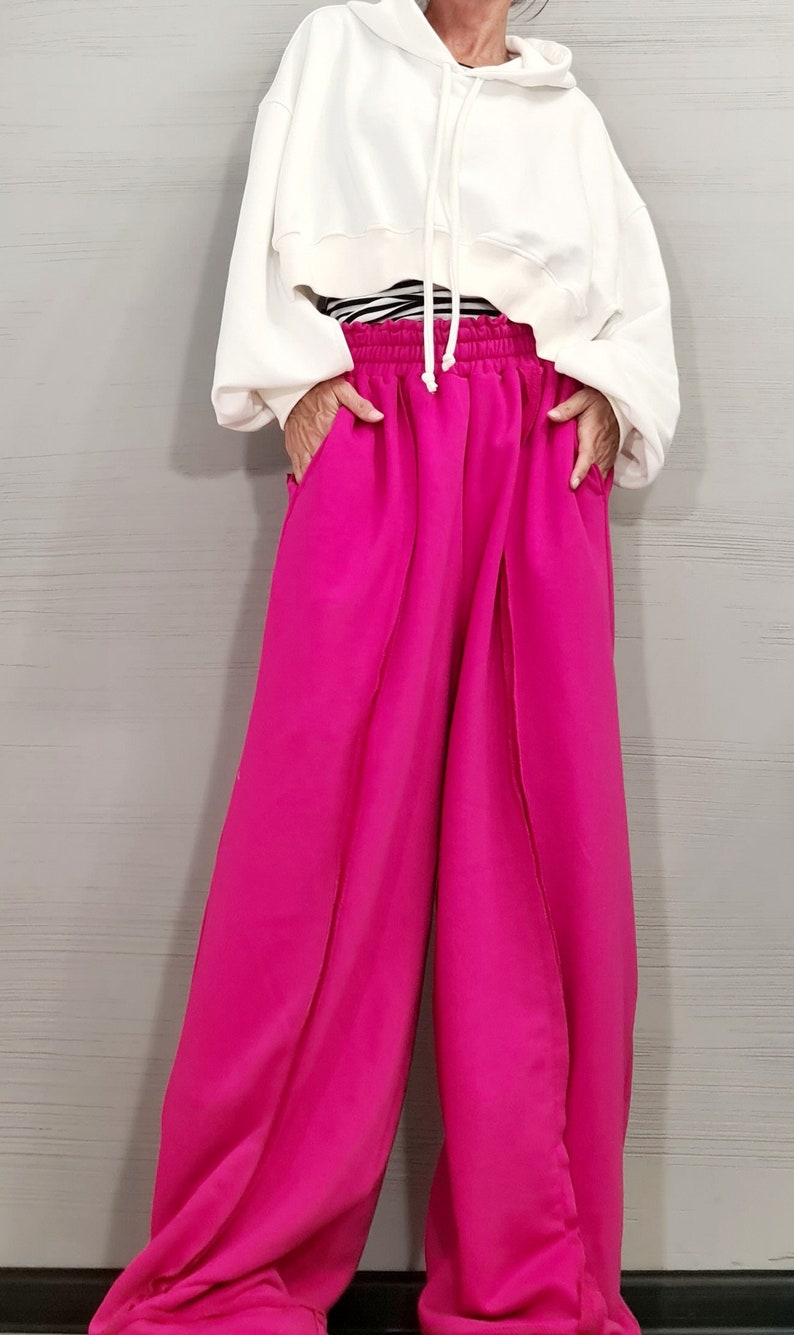 May include: A white hooded sweatshirt with a drawstring is paired with vibrant fuchsia wide-leg pants. The pants have a high waist and pockets. A black and white striped top is visible beneath the sweatshirt.