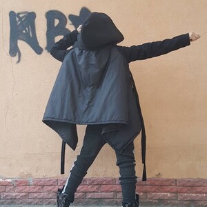 K&ouml;nnte beinhalten: Eine schwarze Kapuzenjacke mit glattem, gestepptem R&uuml;ckenteil und langen &Auml;rmeln, kombiniert mit einer dunkelgrauen, strukturierten Hose mit hellem Seitenstreifen und schwarzen High-Top-Stiefeln mit gl&auml;nzender, gemusterter Oberfl&auml;che.