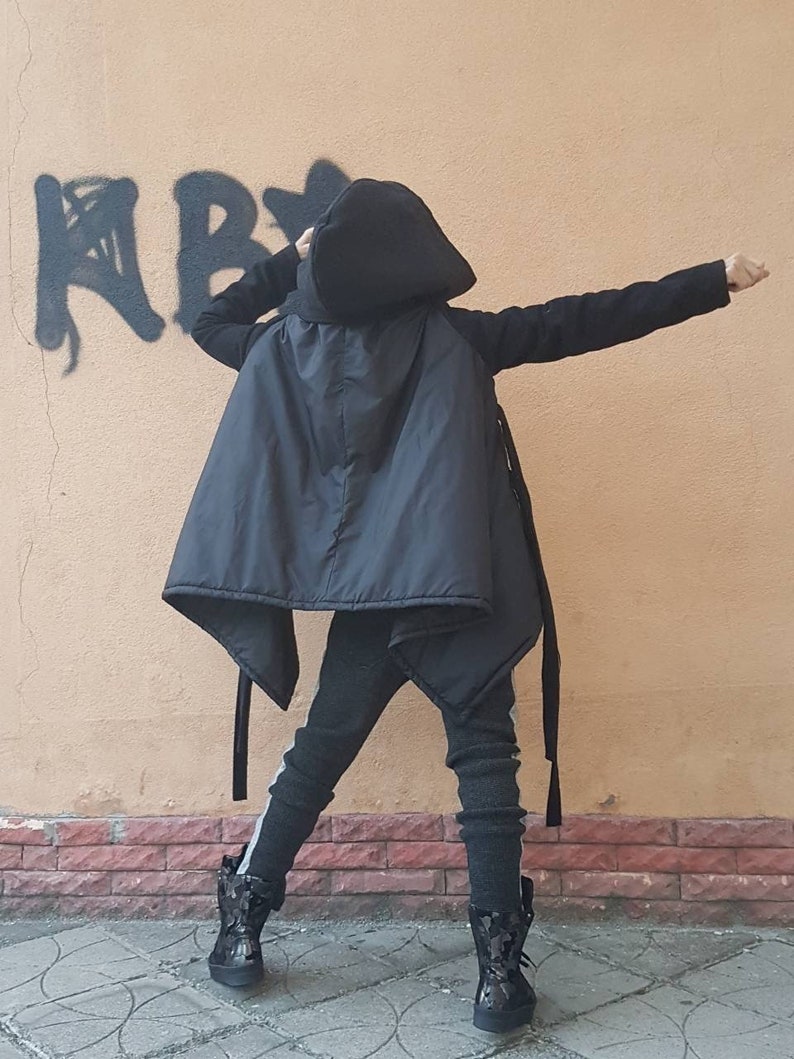 K&ouml;nnte beinhalten: Eine schwarze Kapuzenjacke mit glattem, gestepptem R&uuml;ckenteil und langen &Auml;rmeln, kombiniert mit einer dunkelgrauen, strukturierten Hose mit hellem Seitenstreifen und schwarzen High-Top-Stiefeln mit gl&auml;nzender, gemusterter Oberfl&auml;che.