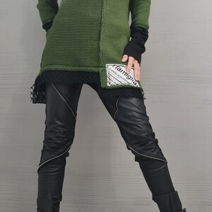 May include: An olive green cold-shoulder knit sweater with a black underlayer, featuring a white patch with the text 'famiglia' on the lower front. Paired with black trousers featuring diagonal silver zippers and black platform boots with buckle straps.
