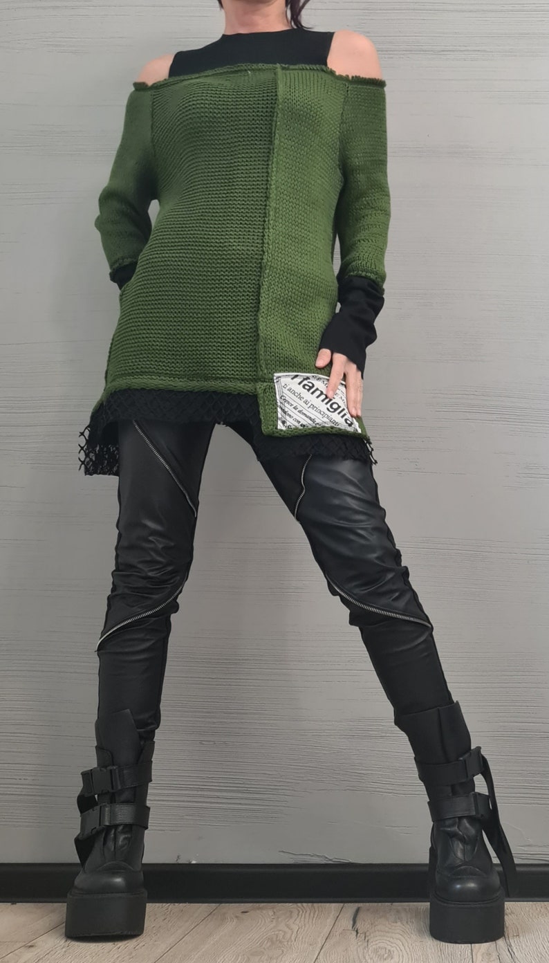 May include: An olive green cold-shoulder knit sweater with a black underlayer, featuring a white patch with the text 'famiglia' on the lower front. Paired with black trousers featuring diagonal silver zippers and black platform boots with buckle straps.