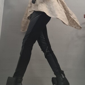 May include: A sleeveless beige turtleneck sweater with a textured knit pattern, paired with black trousers featuring diagonal silver zippers and black leather ankle boots with block heels and small attached pouches on the outer sides.