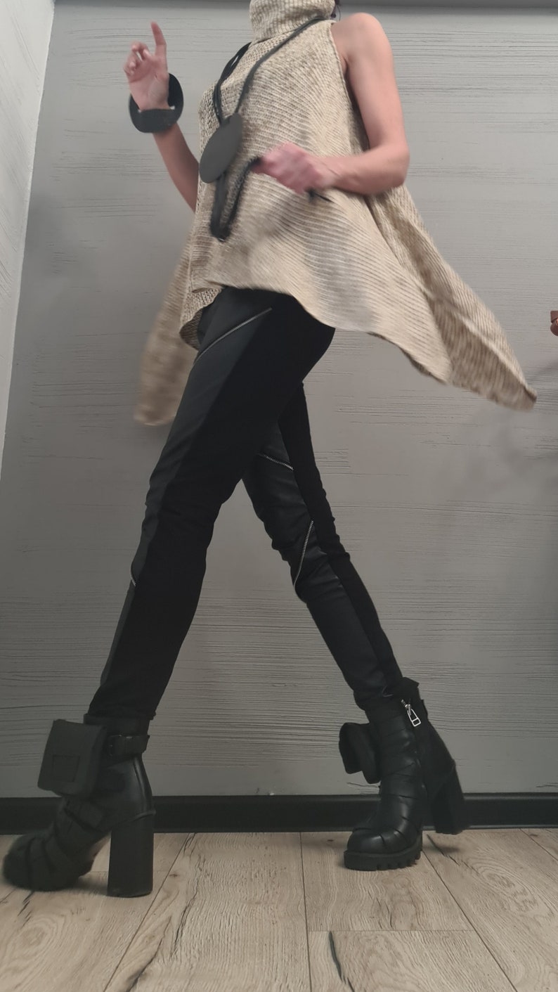 May include: A sleeveless beige turtleneck sweater with a textured knit pattern, paired with black trousers featuring diagonal silver zippers and black leather ankle boots with block heels and small attached pouches on the outer sides.