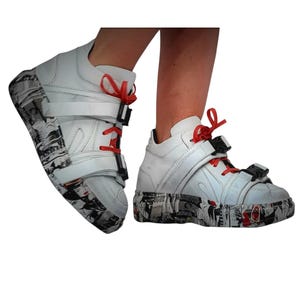Pu&ograve; includere: Sneakers alte bianche con lacci rossi e dettagli neri. Le suole presentano una stampa grafica in bianco e nero. Le scarpe hanno una chiusura con cinturino e sono progettate per uno stile urbano moderno.