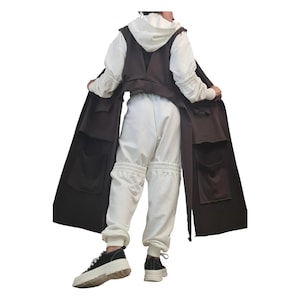 May include: A long, dark brown sleeveless vest featuring large patch pockets, worn over a white hooded sweatshirt and matching white sweatpants with gathered cuffs. The outfit is completed with black and white platform sneakers.