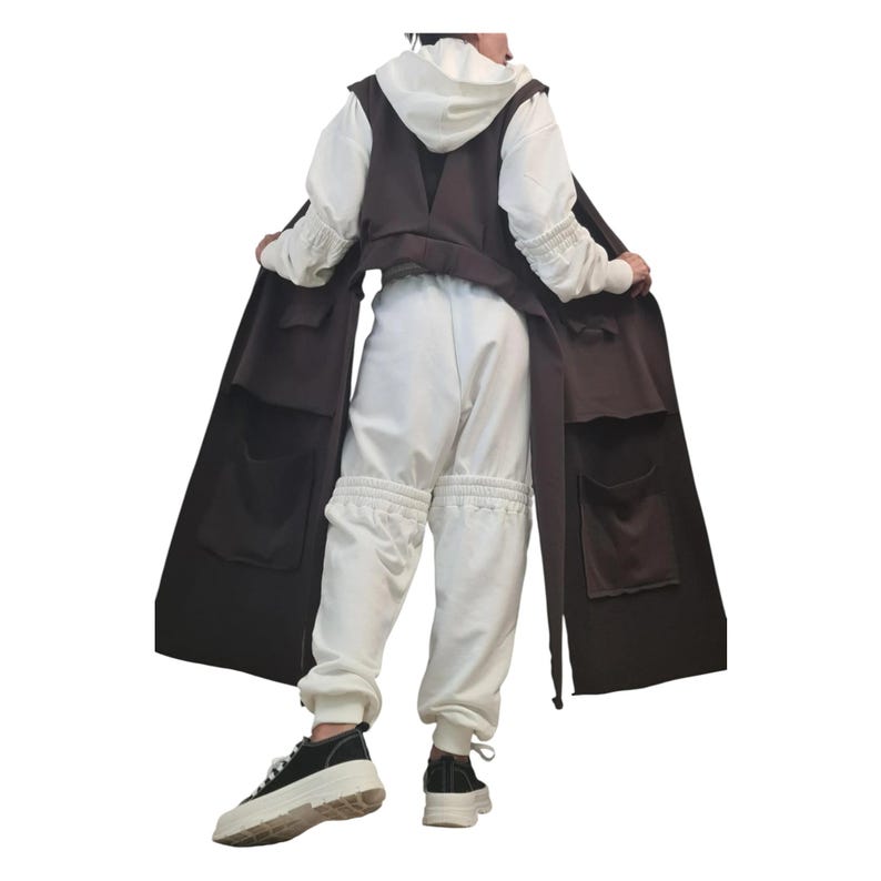 May include: A long, dark brown sleeveless vest featuring large patch pockets, worn over a white hooded sweatshirt and matching white sweatpants with gathered cuffs. The outfit is completed with black and white platform sneakers.