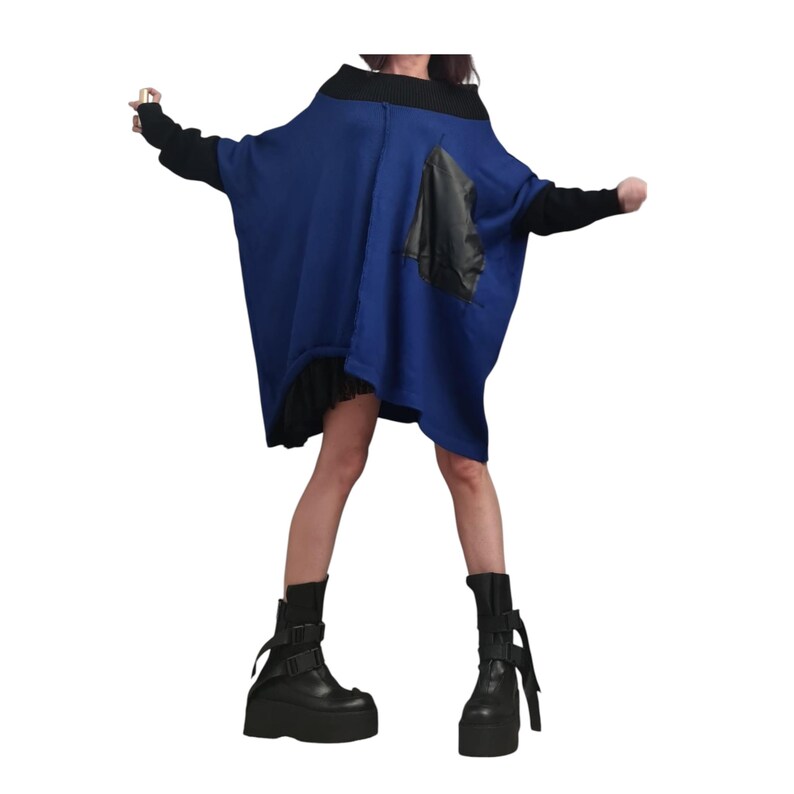 May include: An oversized blue tunic with a black ribbed collar and long black sleeves. The garment features a large black patch pocket on the front and a black pleated skirt hem visible underneath. Black platform boots with buckle straps are worn.