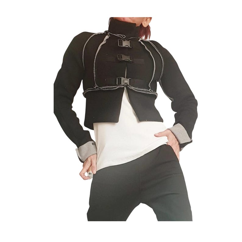 Op de afbeelding: Een zwart jack met lange mouwen, een hoge kraag, drie plastic gespsluitingen aan de voorzijde en zichtbare grijze naaddetails. Het jack heeft grijze boorden en wordt gedragen over een wit shirt in combinatie met een zwarte broek.
