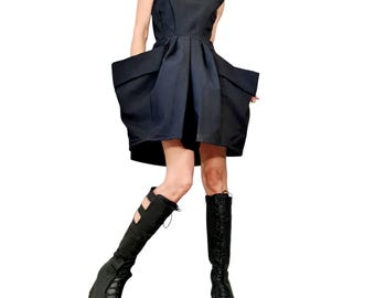 Puede incluir: Vestido negro sin mangas con cuello redondo y bolsillos grandes y estructurados. El vestido se combina con botas de plataforma negras. El estilo general es moderno y atrevido.