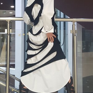 May include: A white shirt with a black, cut-out design, paired with black sandals. The outfit features a unique, layered aesthetic. The model is wearing black sandals.