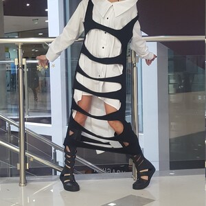 May include: A white button-down shirt is layered under a black, cut-out dress. The dress has a unique design with vertical slits. The outfit is paired with black, open-toe, gladiator-style sandals. The overall look is modern and edgy.