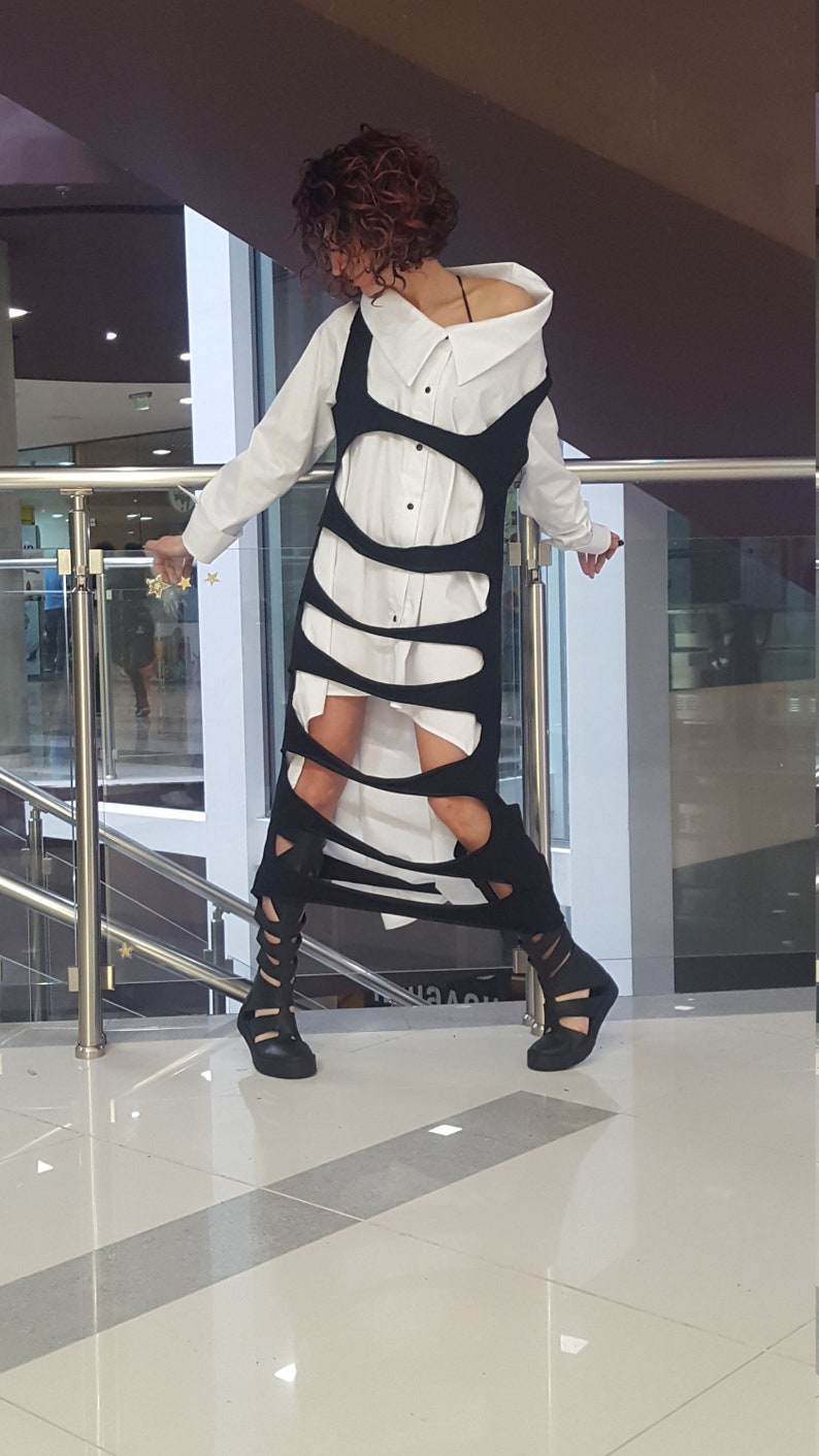 May include: A white button-down shirt is layered under a black, cut-out dress. The dress has a unique design with vertical slits. The outfit is paired with black, open-toe, gladiator-style sandals. The overall look is modern and edgy.