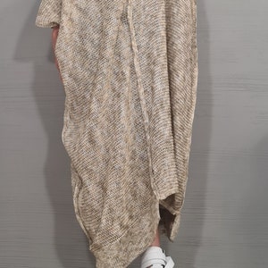 May include: An oversized, loose-fitting beige and white marled knit tunic with short sleeves and a visible vertical seam running down the center. The garment features a wide, asymmetrical neckline and a textured, open-weave fabric construction.