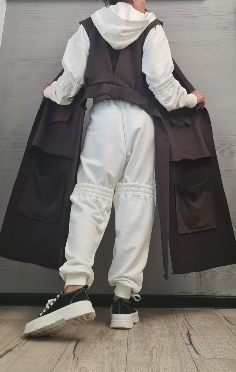 May include: A white hooded sweatshirt with gathered sleeves, white sweatpants featuring gathered knee sections, and a long, sleeveless dark brown vest with large patch pockets. The outfit is paired with black sneakers with thick white soles.