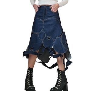 Può includere: Gonna in denim blu scuro con un esclusivo design a cerchi ritagliati. La gonna è abbinata a stivali neri stringati con dettagli a catena argentata. L'outfit è completato da un maglione bianco a coste con spalle scoperte.