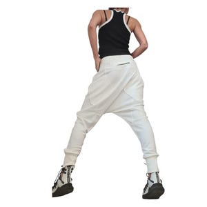 Op de afbeelding: Witte harembroek met een lage kruis en aansluitende enkels. De broek heeft grote zakken en een ritssluiting aan de achterkant. Gecombineerd met een zwarte tanktop met witte bies en zwart-witte sneakers.