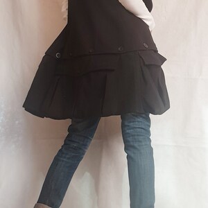 May include: A black vest with a ruffled hem and a white shirt underneath. The vest has a collar and is worn over a white shirt. The person is wearing blue jeans and black boots.
