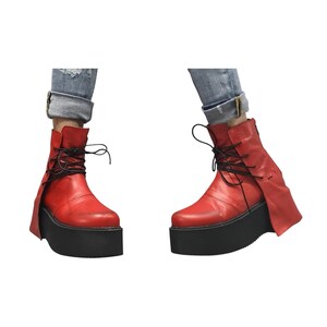 K&ouml;nnte beinhalten: Rote Leder-Ankle Boots mit schwarzen Plateausohlen und schwarzen Schn&uuml;rsenkeln. Die Stiefel haben ein einzigartiges Design mit einer Klappe an der Seite. Die Stiefel werden mit blauen Jeans getragen.