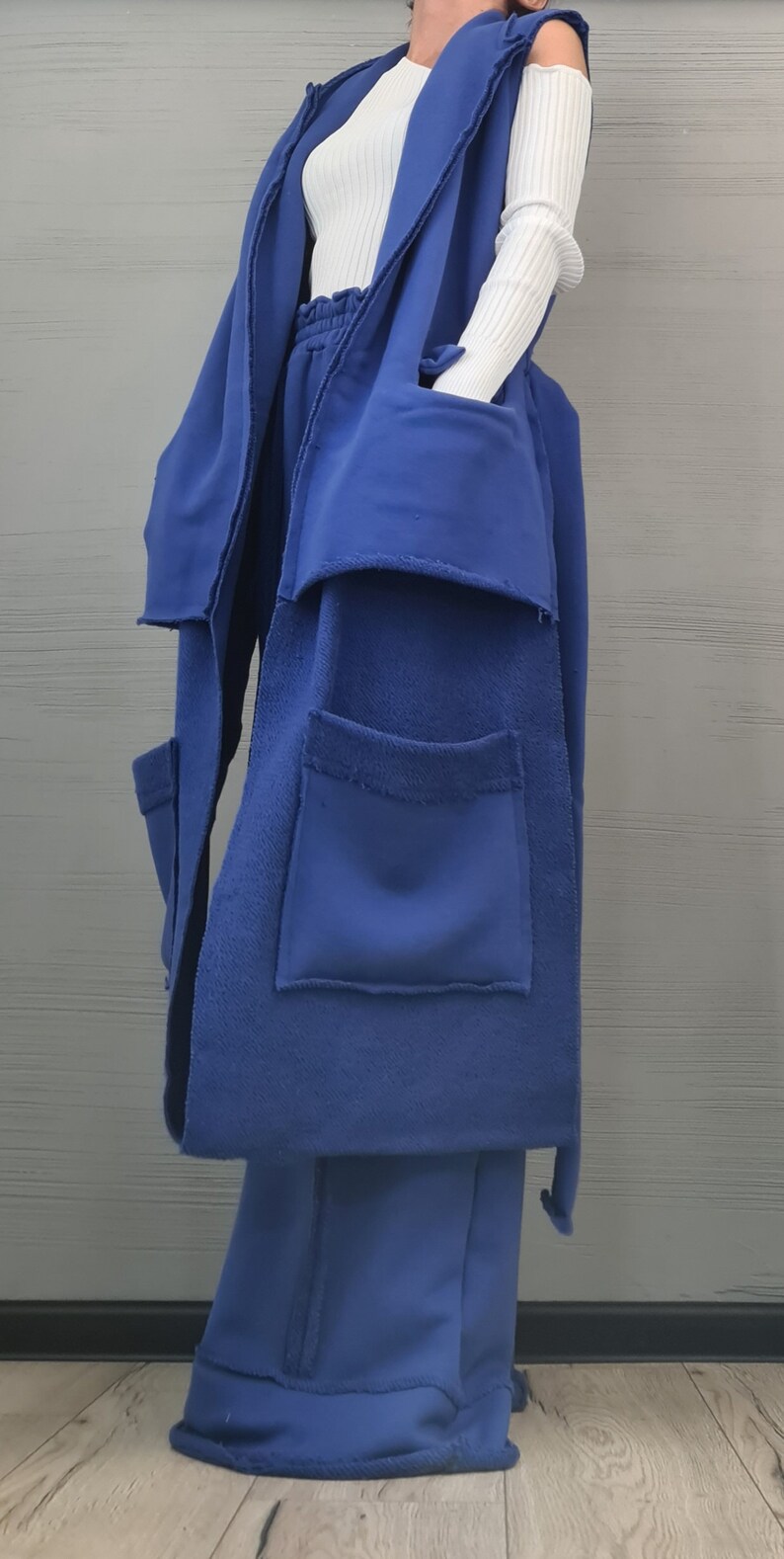 May include: A long, sleeveless royal blue vest with raw, frayed edges and large patch pockets, worn over a white ribbed long-sleeve top and matching royal blue wide-leg trousers with an elasticized waistband and visible seam detailing.
