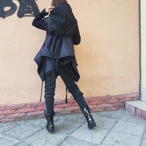 K&ouml;nnte beinhalten: Eine schwarze Kapuzenjacke mit geschichtetem, asymmetrischem Saum und Taillenband, kombiniert mit dunkelgrauer, strukturierter Leggings und schwarzen Stiefeln. Die Jacke hat lange &Auml;rmel und eine gesteppte Textur, die eine geschichtete Silhouette erzeugt.