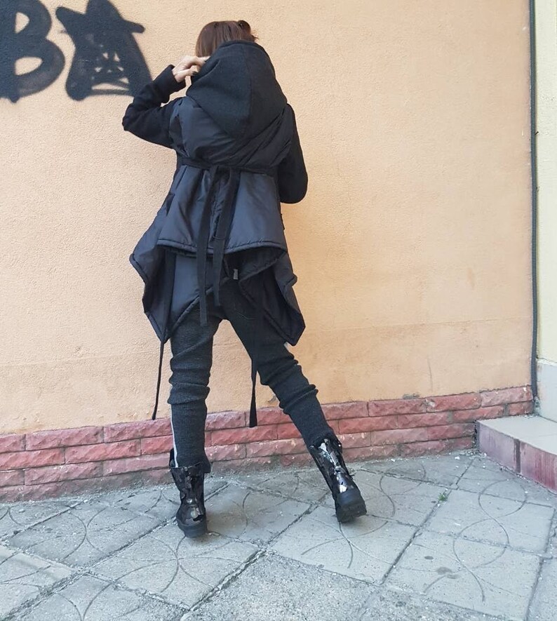 K&ouml;nnte beinhalten: Eine schwarze Kapuzenjacke mit geschichtetem, asymmetrischem Saum und Taillenband, kombiniert mit dunkelgrauer, strukturierter Leggings und schwarzen Stiefeln. Die Jacke hat lange &Auml;rmel und eine gesteppte Textur, die eine geschichtete Silhouette erzeugt.