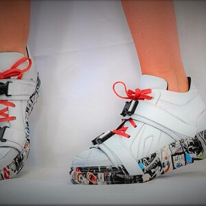 Pu&ograve; includere: Sneakers alte bianche con lacci e dettagli rossi. Le scarpe presentano fibbie nere e una suola fantasia con un collage di immagini. Design moderno e audace.