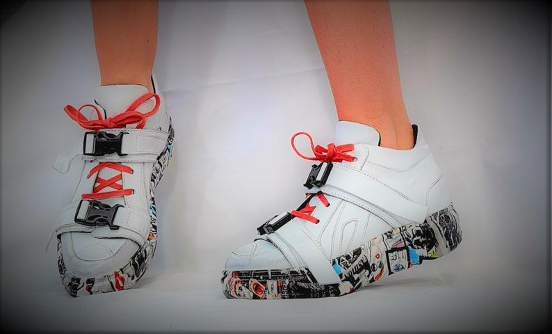 Pu&ograve; includere: Sneakers alte bianche con lacci e dettagli rossi. Le scarpe presentano fibbie nere e una suola fantasia con un collage di immagini. Design moderno e audace.
