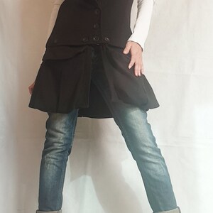 May include: A long black sleeveless vest with a large collar and a skirt-like bottom. The vest has a button closure and a belt. The person is wearing blue jeans and black boots.