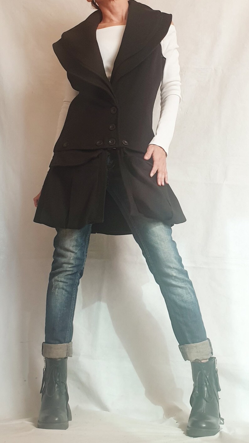 May include: A long black sleeveless vest with a large collar and a skirt-like bottom. The vest has a button closure and a belt. The person is wearing blue jeans and black boots.