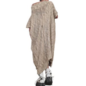 May include: A loose-fitting, beige and tan heathered knit tunic dress with short sleeves and a visible vertical seam running down the center. The garment features an open, textured weave and a wide, relaxed neckline that drapes off the shoulder.