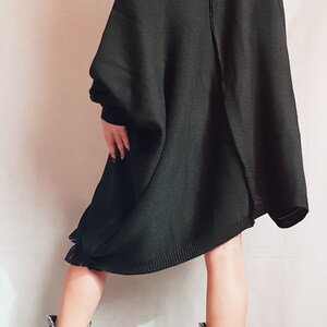 May include: An oversized black knit sweater with a wide, off-the-shoulder neckline and a visible vertical seam down the back. The sweater features long sleeves and a loose, flowing silhouette. Paired with black boots featuring white graphic text.