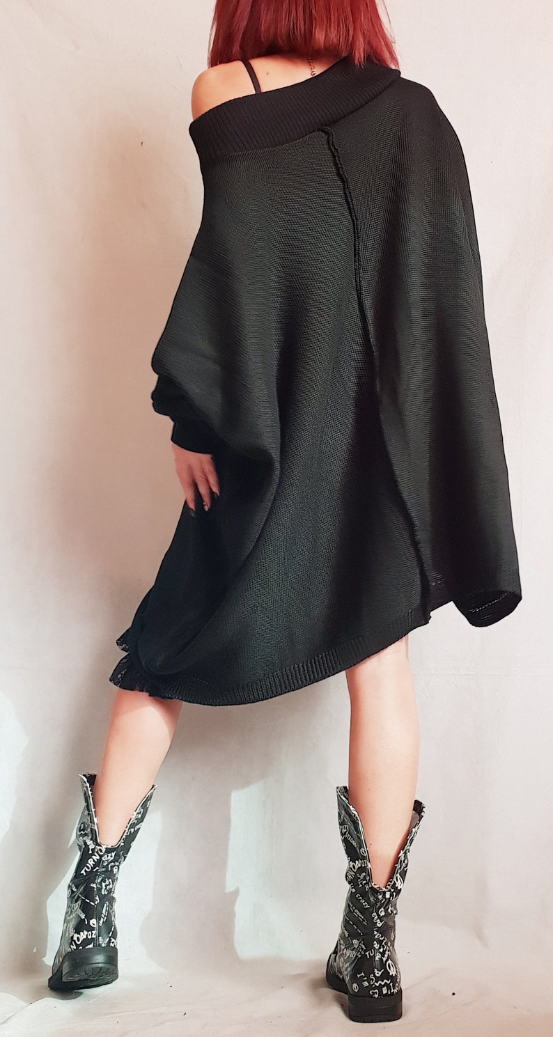 May include: An oversized black knit sweater with a wide, off-the-shoulder neckline and a visible vertical seam down the back. The sweater features long sleeves and a loose, flowing silhouette. Paired with black boots featuring white graphic text.