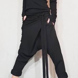 May include: Black ensemble featuring a turtleneck top with shoulder cutouts and long sleeves. The outfit includes draped pants with a long tie and black ankle boots. The pants have pockets and the cuffs are rolled up.