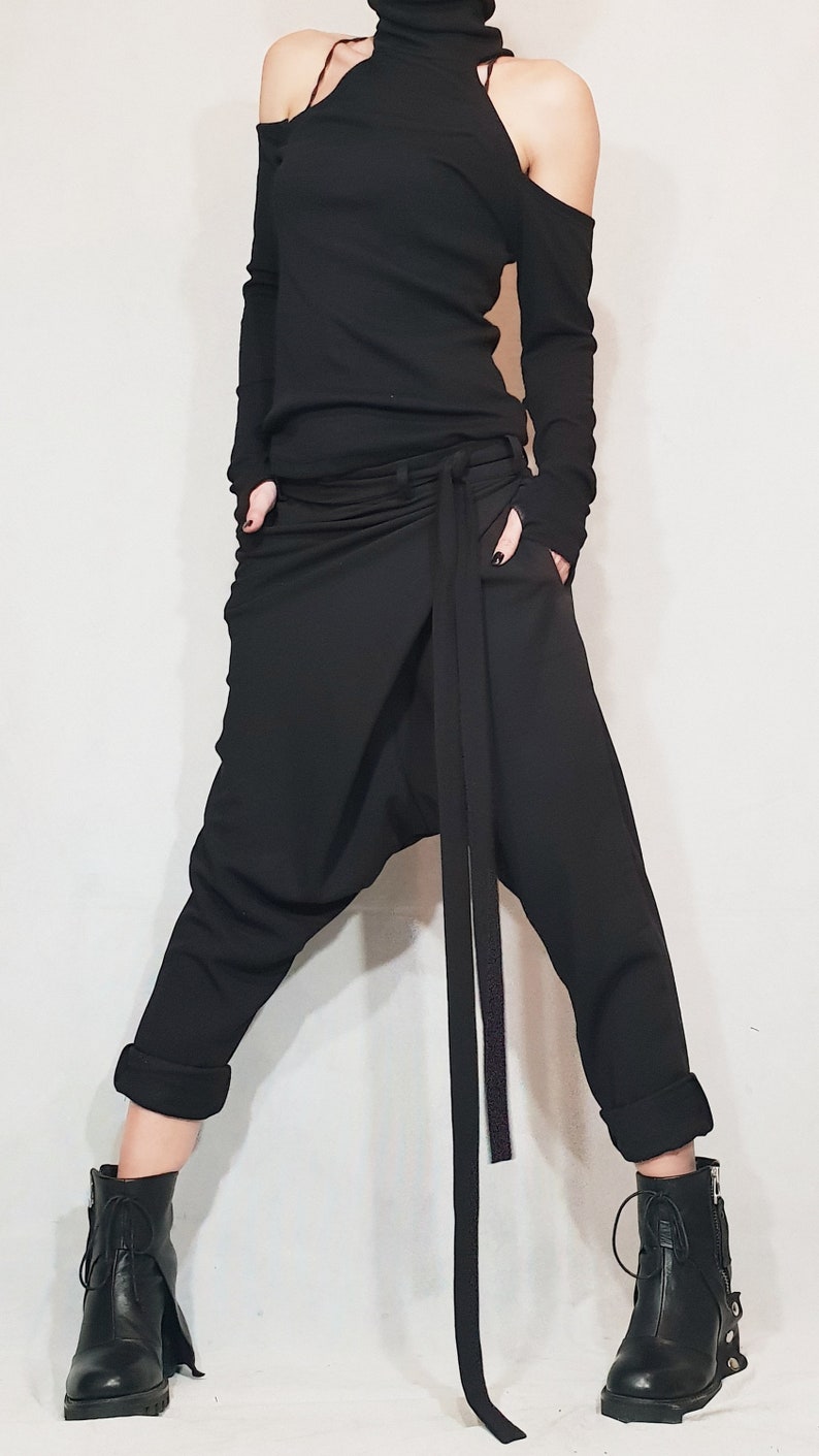 May include: Black ensemble featuring a turtleneck top with shoulder cutouts and long sleeves. The outfit includes draped pants with a long tie and black ankle boots. The pants have pockets and the cuffs are rolled up.