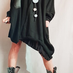 May include: An oversized black knit tunic with an asymmetrical hem, a ribbed off-the-shoulder neckline, and a black faux leather patch on the right sleeve. It is paired with a long necklace featuring circular pendants and black patterned boots.