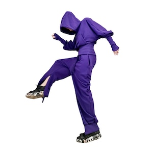 May include: A purple hooded sweatshirt and matching sweatpants set. The sweatshirt has a unique, oversized design with a hood and long sleeves. The sweatpants have a side slit. The person is wearing black and white patterned shoes.