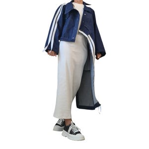 May include: A dark blue denim jacket with white stripes on the sleeves and a long, asymmetrical design. The outfit includes a white top, white pants, and black and white sneakers.