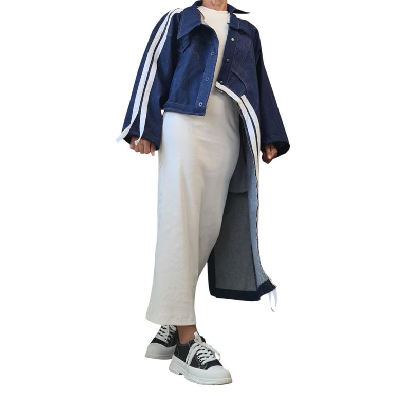 May include: A dark blue denim jacket with white stripes on the sleeves and a long, asymmetrical design. The outfit includes a white top, white pants, and black and white sneakers.