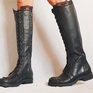May include: Black knee-high boots with a lace-up design and studded toe caps. The boots have a zipper on the side and are worn with black lace trim at the top. The boots are made of leather and have a flat sole.
