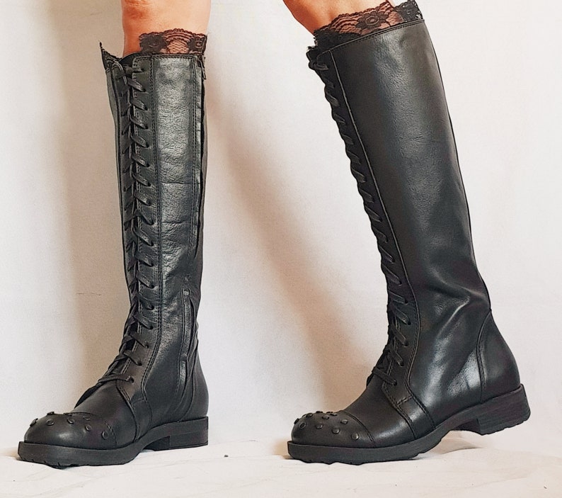 May include: Black knee-high boots with a lace-up design and studded toe caps. The boots have a zipper on the side and are worn with black lace trim at the top. The boots are made of leather and have a flat sole.