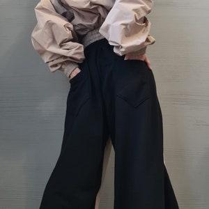 May include: A beige hooded windbreaker jacket with long sleeves and a ribbed waistband paired with wide-leg black trousers featuring two back pockets. The jacket has a loose, voluminous fit, while the trousers have a relaxed, flowing silhouette.