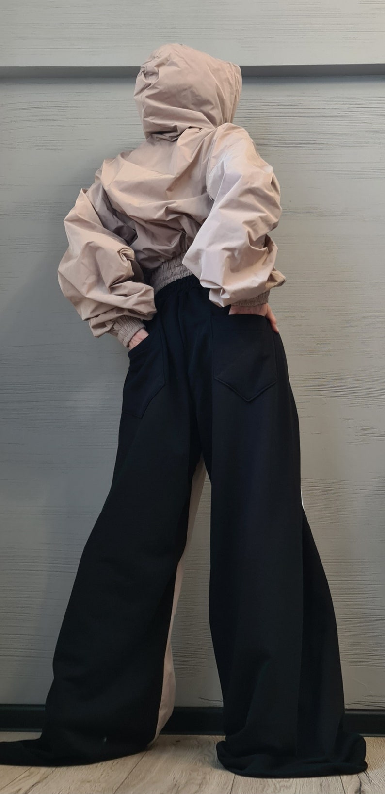 May include: A beige hooded windbreaker jacket with long sleeves and a ribbed waistband paired with wide-leg black trousers featuring two back pockets. The jacket has a loose, voluminous fit, while the trousers have a relaxed, flowing silhouette.