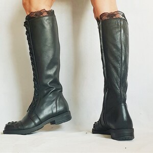 May include: Black knee-high boots with a lace-up front and a zipper on the side. The boots have a square toe and are made of a smooth, dark material. The top of the boots are trimmed with black lace.